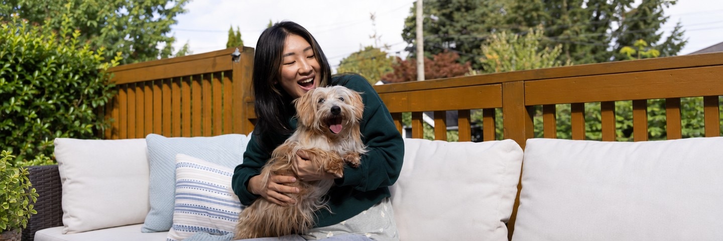 야외 소파에 앉아 갈색 털의 강아지를 안고 웃는 보호자