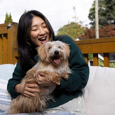 야외 소파에 앉아 갈색 털의 강아지를 안고 웃는 보호자